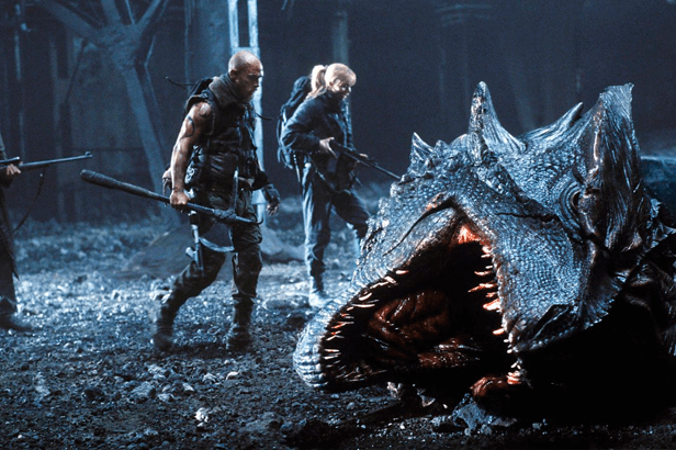 Cena do filme Reino de Fogo com um homem e uma mulher em pé ao lado da cabeça de um dragão